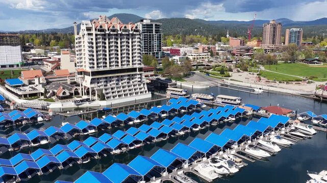 Coeur d'Alene Resort and marina with blue-roofed boat slips on Coeur d'Alene Lake, showing waterfront architecture and mountain landscape in Idaho, United States. Aerial flyover.