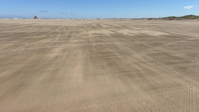 Static view of wind-blown sand flowing across a flat sandy beach near the ocean.