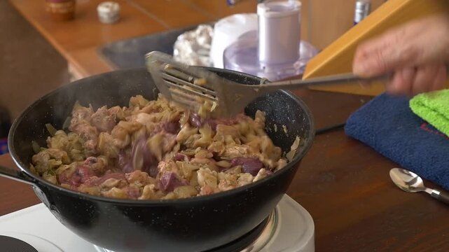 Close-up of a hand using a spatula to stir partridge meat and saut&eacute;ed onions in a black pan.