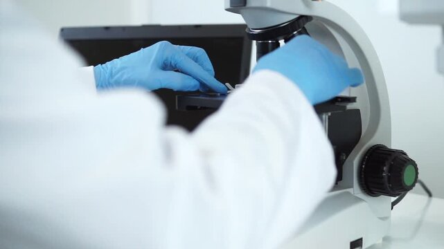 A scientist in a white lab coat and blue gloves carefully places a glass slide with a blood sample onto a modern microscope for Hantavirus analysis.