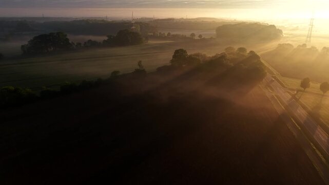 Morning traffic at rural road intersection in United States, with sun rays piercing through thick fog and trees during golden sunrise. Aerial drone flyover shot. Wide shot. Peaceful countryside scene.