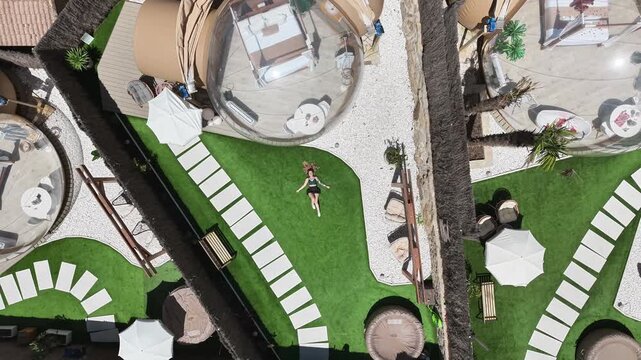 Aerial drone shot rising above a luxury glamping resort in Cazorla, Spain, with a woman relaxing on a green lawn beside dome accommodation and spa area.