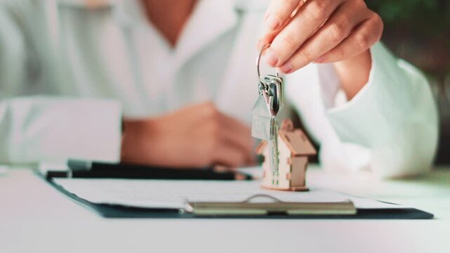 Hand presenting keys with house shaped keychain above clipboard contract. Concept of buying first home, mortgage paperwork, real estate transaction and future property ownership success.