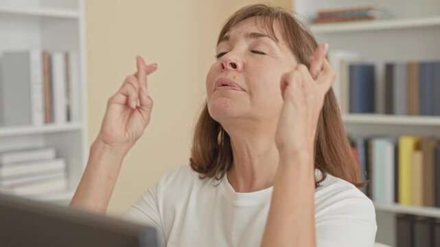 Woman crosses fingers at computer in building office, eyes closed and hands raised in a quiet prayer pose; hope.