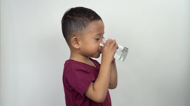 A 4 year old Asian boy is thirsty and drinks water from a transparent glass.