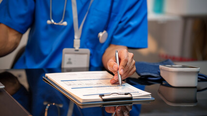 Close-up of healthcare worker in blue scrubs filling out medical forms on clipboard at desk, with...