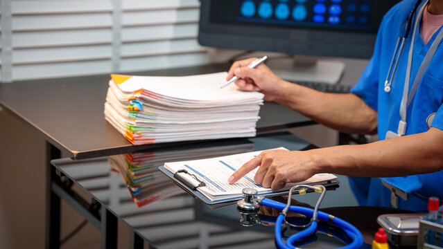 Healthcare worker at desk reviewing patient paperwork and clipboard with stethoscope, hospital monitor in background, medical administration and documentation concept.