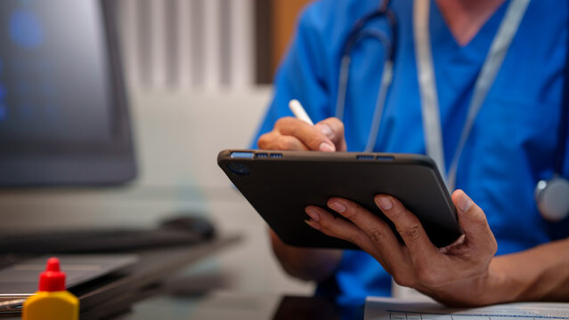 Close-up of heaalthcare worker in blue scrubs writing on a digital tablet with stylus at desk, stethoscope visible, computer monitor blurred in background, modern clinic office.
