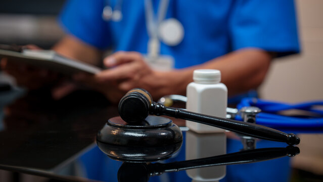 Medical malpractice legal concept with judge gavel on desk, prescription bottle and stethoscope, doctor in scrubs holding clipboard in background.