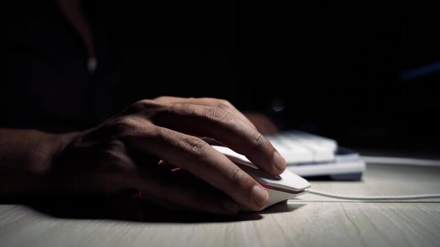 Close-up of a person&rsquo;s hand using a white computer mouse on a desk in a dark room