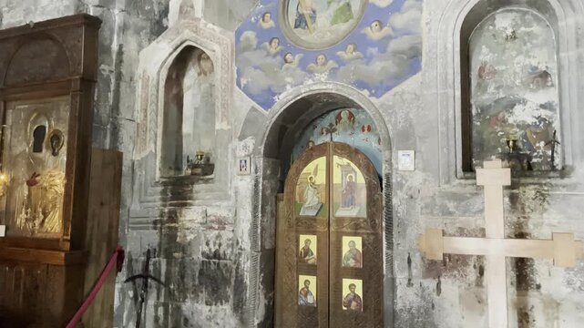 Interior view of the ancient Ananuri Fortress church in Georgia, showing the ornate wooden Royal Doors of the iconostasis with painted saints, set against weathered stone walls