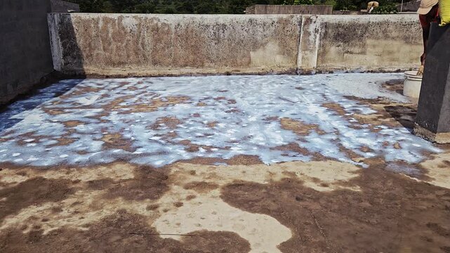 Waterproofing a concrete terrace floor by applying a white URP liquid coating. A worker uses a broom and a bucket to spread the sealant over the damp, stained surface near a textured gray wall.