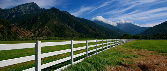 Fototapeta premium White fence green field mountain landscape blue sky white cloud countryside rural meadow valley scenic view pasture grass nature outdoor farmland