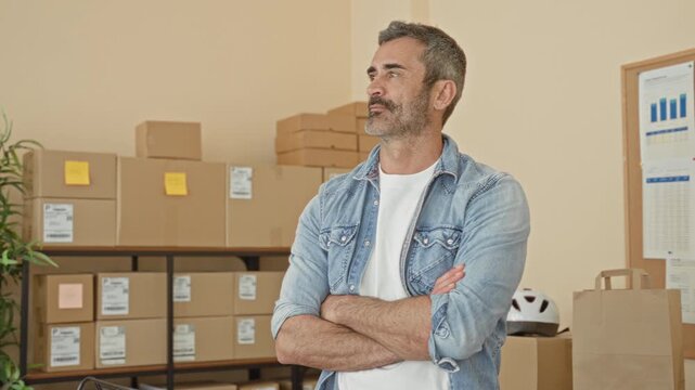 Man with crossed arms and stubble stands beside stacked cardboard boxes, shipping labels, a plant and a helmet in a building; confidence entrepreneurship.