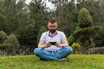 Obraz premium Man sitting cross-legged on green grass, focused on a smartphone in his hands, surrounded by a landscaped garden featuring neatly trimmed shrubs and trees in the background