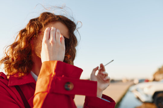 Young woman with curly red hair in a red coat covers her face with one hand while holding a cigarette outdoors on a sunny day near a parking area.