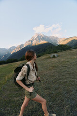 Obraz premium Hiking woman with a backpack strides through a grassy meadow toward distant mountains, focused and determined in a natural outdoor setting, her hair tied back, soft light highlighting the landscape