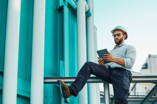 a man in work attire and a helmet is holding a tablet standing next to the side of a very tall blue-green crane with white pipes., quality control