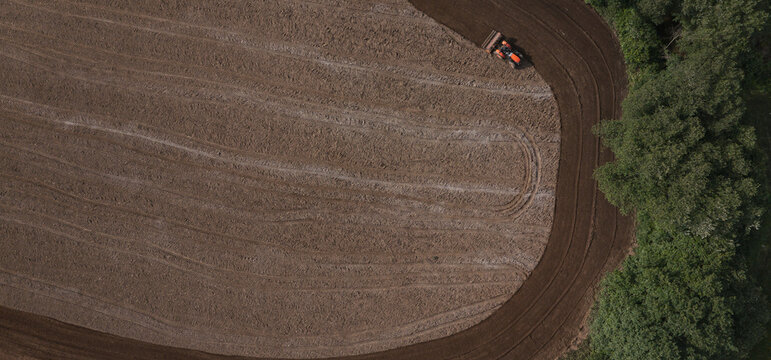 Aerial view of a tractor plowing a brown agricultural field with curved soil patterns next to a green forest edge in Lisbon, Lisbon, Portugal.