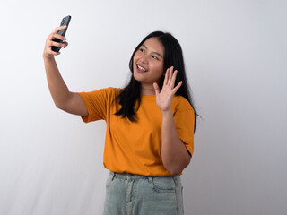 Smiling Asian woman in yellow t-shirt doing video call pose while waving palm saying hi isolated over white background. © Luqman Aditiamala