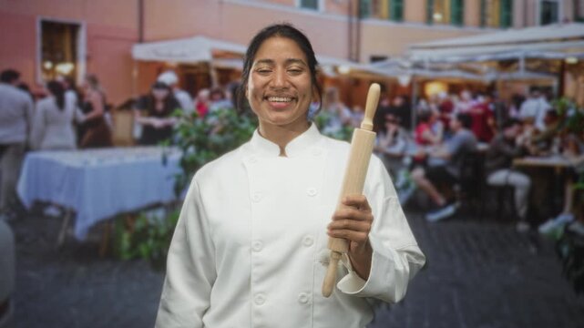 Woman chef smiling with hand holding a rolling pin in a bustling street cafe among outdoor diners and tables; pride hospitality.
