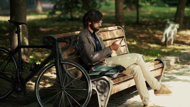 Businessman Browsing Smartphone while Resting on Bench in City Park. Freelancer Working Remotely with Mobile Phone near Bicycle
