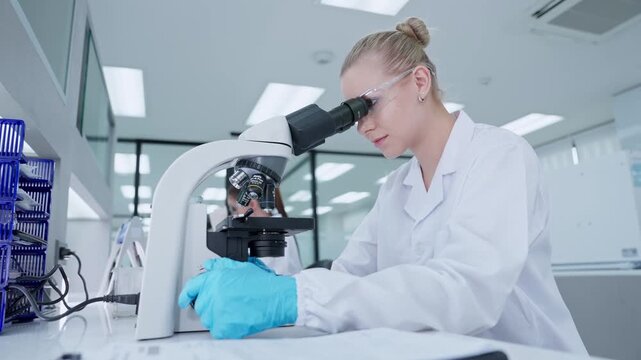 A scientist in a laboratory examines a test tube while using a microscope. Another researcher is working nearby. The focus is on the process of scientific research and discovery.