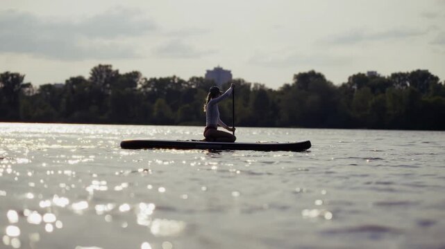 Woman Kneeling on Stand Up Paddleboard and Rowing on River. Training on SUP Board at Sunset. Summer Water Sports and  Fitness