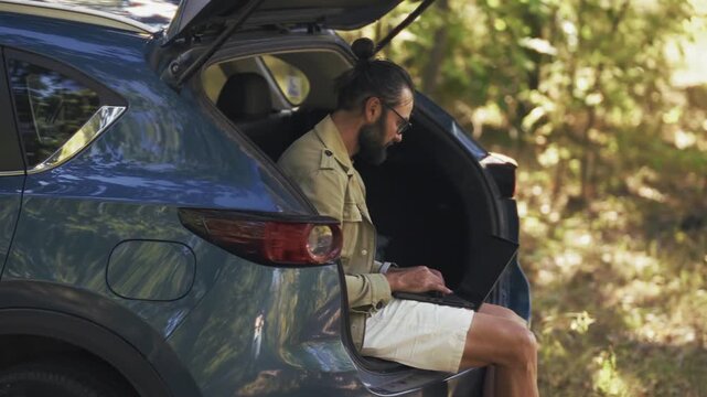 Businessman Working on Laptop while Sitting in Open Trunk of Car. Freelancer Managing Business Tasks During Summer Travel Trip. Digital Nomad Lifestyle