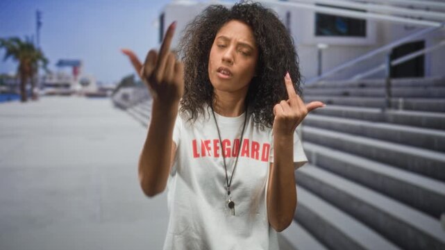 Woman lifeguard with curly hair flips both middle fingers with raised hands on seaside boardwalk steps; defiance.