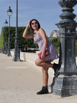 A woman wearing a purple floral sundress, sunglasses, and black lace-up boots leans against a decorative lamppost