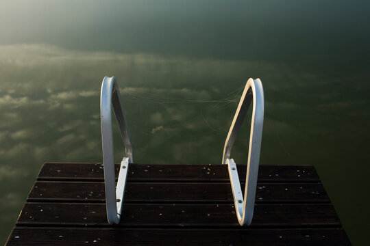 Metal swimming ladder handrails on a wooden lake dock in misty morning light