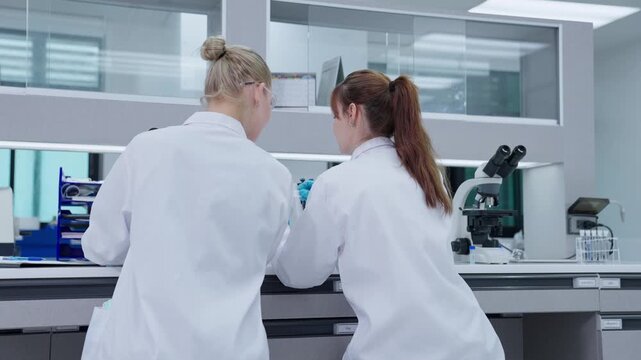 Two scientists in lab coats focus on their work at a modern laboratory. They handle test samples and use equipment on lab benches as they conduct important research.