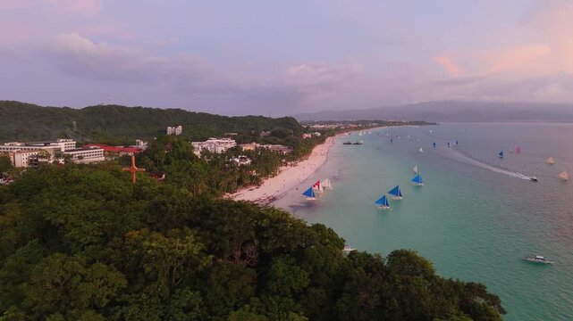 Aerial reveal of Boracay White Beach at sunset showing iconic paraw sailboats on turquoise water, famous cross landmark, resort hotels, and curved white sand shoreline, Philippines