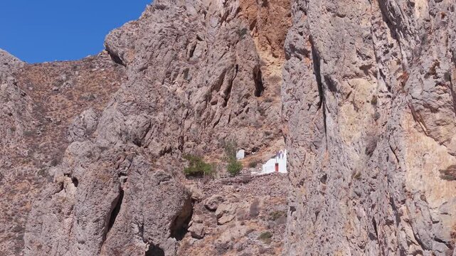 Drone shot revealing a chapel located in the rocks above Perissa Beach in Santorini, showcasing dramatic volcanic cliffs and the stunning Aegean coastal landscape.
