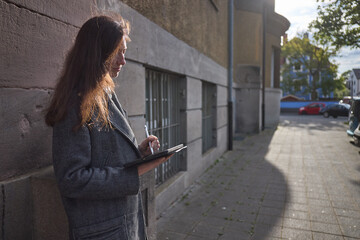 Woman sketching on a tablet device while leaning against a textured wall outdoors