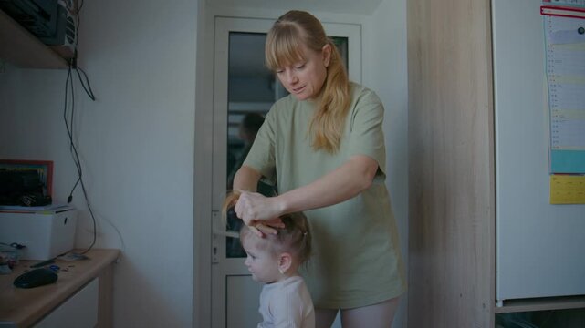 Mother gently brushes her child's hair in a warm, domestic setting. She smiles softly, showing care and affection. Child stands calmly, trusting her mother's touch