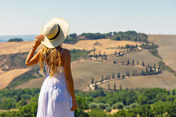 Fototapeta premium Back view of woman in white dress holding straw hat looking at cypress winding road in Val d'Orcia Tuscany. travel destination, summer vacation in Italy