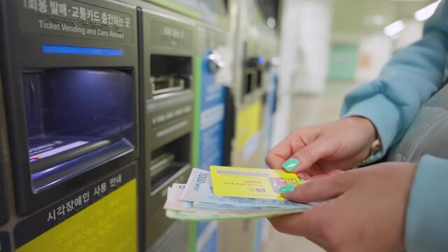 Woman with turquoise nail polish purchases metro ticket at automated vending machine in Seoul South Korea subway station. Hands holding transit card and Korean won cash bills at self-service kiosk.