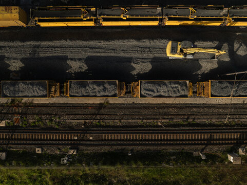 Aerial view of a yellow excavator working near freight train wagons filled with gravel on railway tracks in Priolo Gargallo, Sicily, Italy.