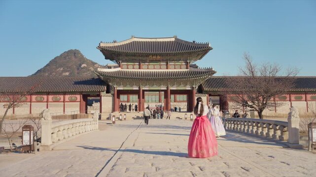 Tourists wearing traditional Korean hanbok dresses walk toward ornate gate at Gyeongbokgung Palace in Seoul, South Korea. Clear blue sky frames the historic royal architecture with mountain backdrop.