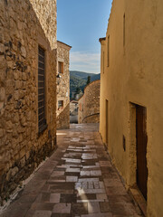Fototapeta premium Empty narrow alley between old stone and plaster houses in Mediterranean village during warm sunlight. Historic street with textured walls, stone pavement and peaceful atmosphere.