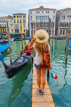 Back view of woman with backpack holding Italian flag on a wooden pier in front of gondolas and palaces in Venice, Italy