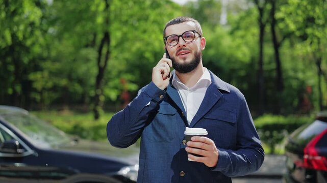 Confident businessman, enjoying coffee, converses happily on smartphone. Environment of green trees and urban setting adds to relaxed, positive mood.