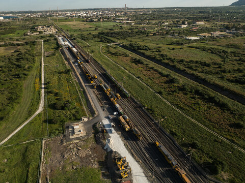 Aerial view of railway maintenance trains on tracks surrounded by green fields and industrial infrastructure in Priolo Gargallo, Sicily, Italy.