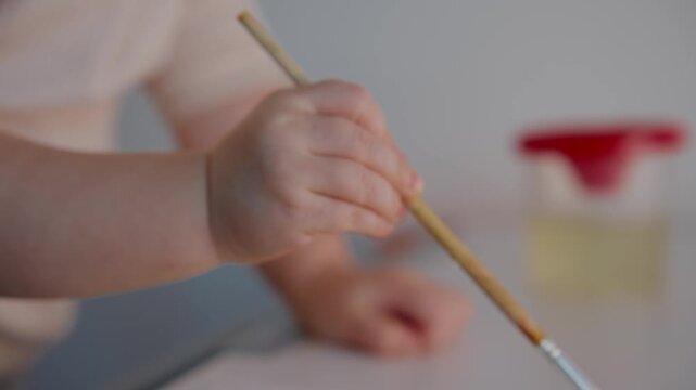Child's small hands hold paintbrush with care. Focused on art, tiny fingers grip brush firmly. Colorful drawing unfolds on paper surface. Background shows art supplies softly blurred