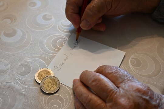 Close-up of an elderly woman's hand writing a shopping list next to a few euro coins, representing budgeting, cost of living, and daily life, Copy Space for text.