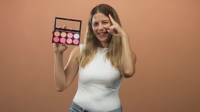 Woman holding a makeup palette with mirror, smiling and pointing finger to temple in studio peach backdrop; cheerful confidence.
