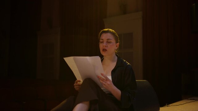 Thoughtful young performer rehearsing lines alone on dimly lit theater stage, sitting near speaker and reading script pages in an intimate dramatic moment