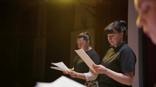 Actors rehearsing dramatic script on dimly lit stage, reading dialogue from printed pages during an intimate theater performance practice session